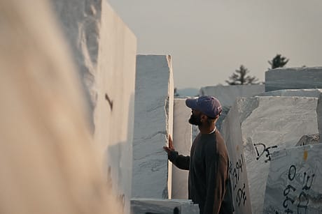 Man wearing a cap examining large marble blocks in a quarry with trees in the background.