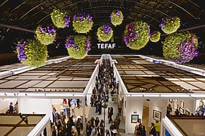 Art fair with hanging floral displays and crowded aisles under a high ceiling, TEFAF sign visible in background.