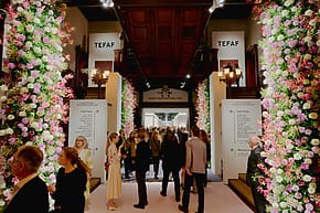 People walking through a flower-decorated entrance at an art fair with signs overhead and flower arrangements along the sides.