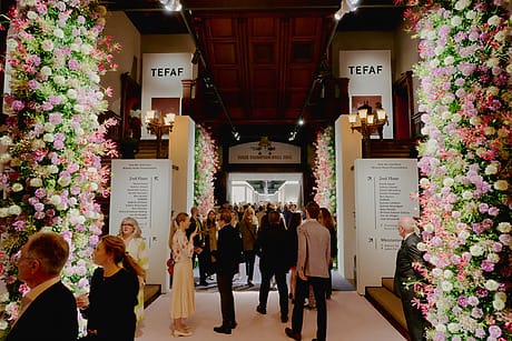 People walking through a flower-decorated entrance at an art fair with signs overhead and flower arrangements along the sides.