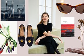Woman seated on a couch surrounded by various artistic items including glasses, shoes, sunglasses, and a painting.