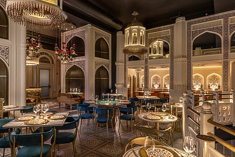 Elegant restaurant interior with ornate decor, chandeliers, patterned tables, blue chairs, and softly lit ambiance.