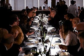 People dining at an elegant long table with dim lighting and glasses of wine.