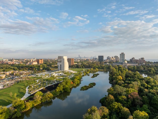 Aerial view of a cityscape with a river, surrounded by lush greenery and buildings under a partly cloudy sky.