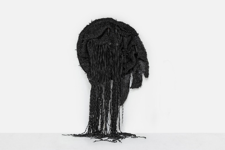 Abstract black textile sculpture with intricate textures and hanging strands displayed against a plain white background.