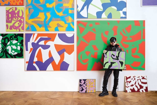 Artist in black attire holding abstract painting, standing in front of large colorful abstract artworks on a gallery wall.