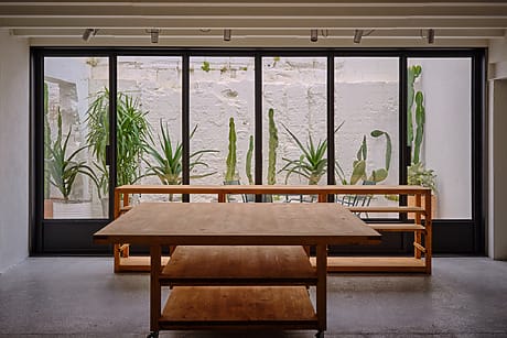Indoor dining area with large wooden tables and chairs, framed by tall potted cacti against large glass windows.