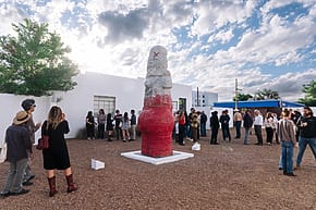 People gathered around a large red and white outdoor sculpture on a sunny day with clouds in the sky.