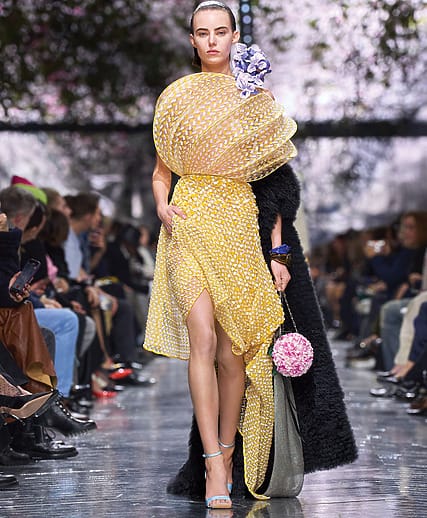 Fashion model walking runway in a vibrant yellow dress with floral patterns, surrounded by audience, carrying a handbag.