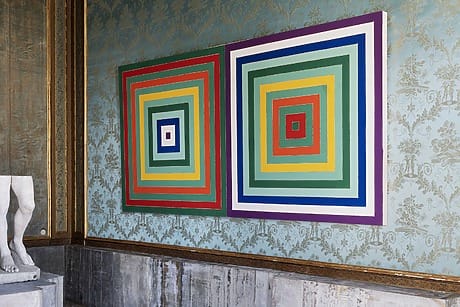 Colorful geometric artwork with concentric squares hangs on an ornate wall next to a partial view of a marble statue.