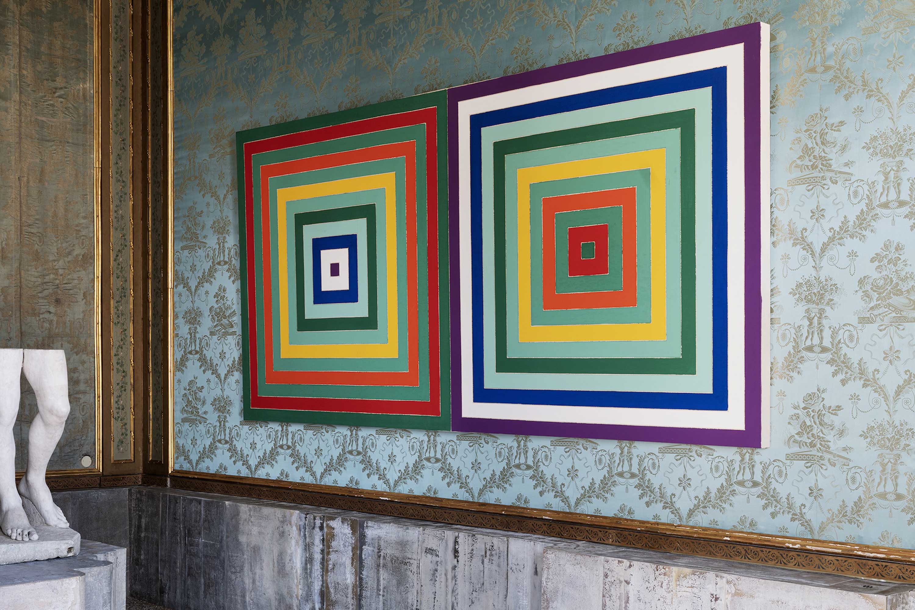 Colorful geometric artwork with concentric squares hangs on an ornate wall next to a partial view of a marble statue.