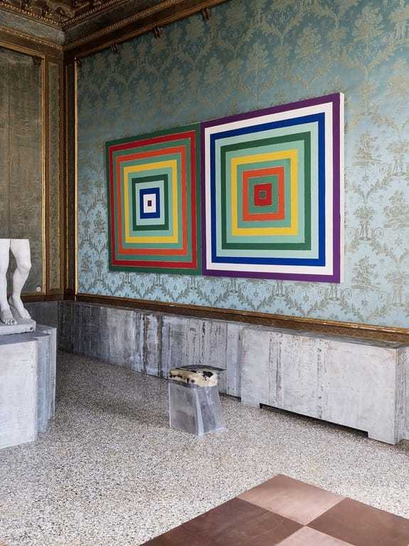 Colorful geometric artwork with concentric squares on ornate wall, statue partial view on the left, modern and classic blend.