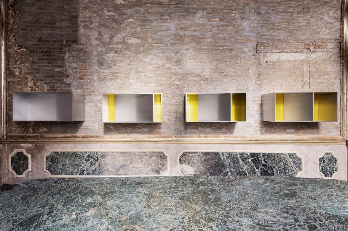 Four modern metal and yellow art installations on a rustic brick wall above a textured marble surface in a gallery setting.