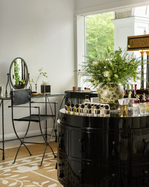 Elegant vanity area with a glossy black cabinet, round mirror, flowers, and perfumes, near a large window with natural light.