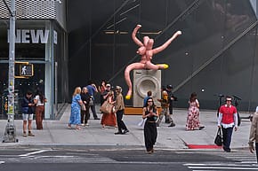 People walking near a modern building with a large, abstract sculpture on the sidewalk.