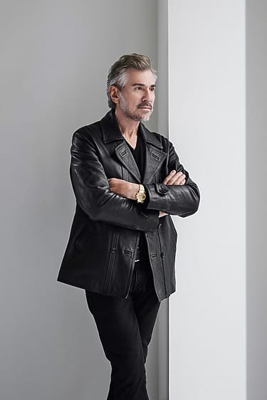 Man in a black leather jacket with arms crossed, standing by a white wall, looking thoughtful.
