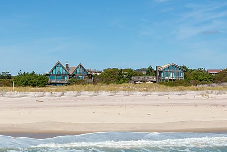 Beachfront view with two modern houses in Fire Island property belonging Michael Kors among dunes and trees under a clear blue sky.