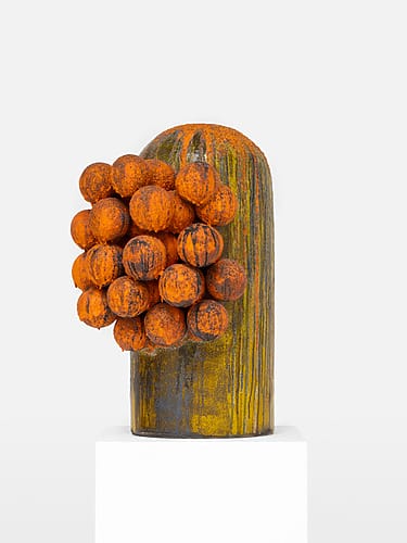 Abstract sculpture with orange textured spheres attached to a tall, cylindrical, rust-hued structure on a white pedestal.