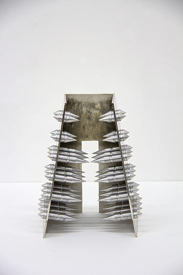Metallic abstract sculpture resembling a tower with conical spikes arranged symmetrically on both sides against a plain background.