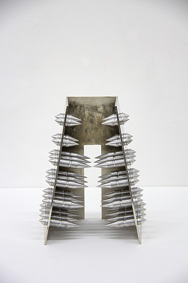 Metallic abstract sculpture resembling a tower with conical spikes arranged symmetrically on both sides against a plain background.