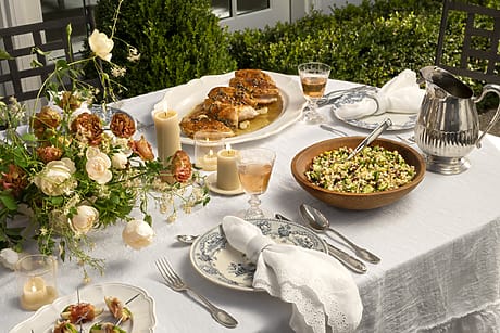 Elegant outdoor dining table with floral centerpiece, roasted chicken, salad, candles, and wine glasses.