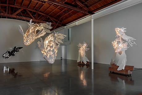 Futuristic art installation with illuminated, white abstract sculptures suspended in a gallery with a wooden ceiling.