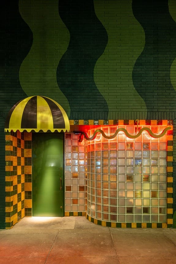 Exterior of a building with green-striped awning, checkered walls, and illuminated glass bricks at night.