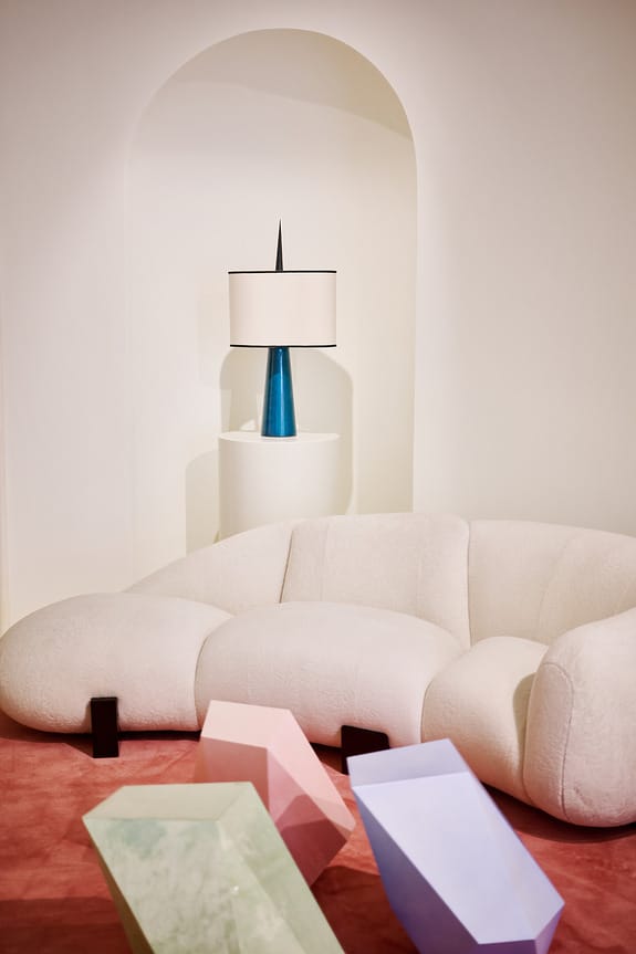 Modern living room with a white curved sofa, geometric tables, and a blue-base lamp on a pedestal against an arched wall.