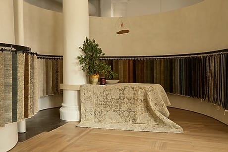Display of various patterned rugs hanging in a showroom with a plant and decorative items on a central table.