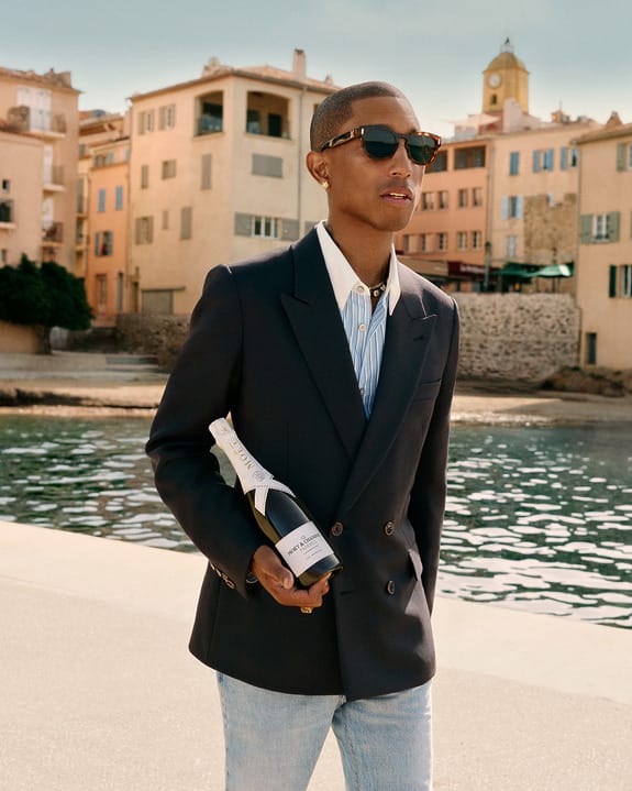Person in sunglasses and suit holding a wine bottle, walking along a waterfront with colorful buildings in the background.